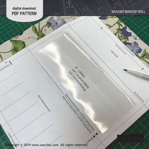Pu&ograve; includere: Un modello PDF digitale per un astuccio per il trucco. Il modello include istruzioni e diagrammi per tagliare e cucire il rotolo. L'immagine mostra i pezzi del modello con etichette di testo, una sovrapposizione trasparente e una penna. Lo sfondo presenta tessuto floreale e un tappetino da taglio verde.