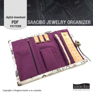 May include: A purple jewelry organizer with multiple compartments and a zipper closure. The organizer is open and shows the interior, which is lined with a white fabric with a black and white print. The organizer has a brown leather strap with metal snaps and a white lace trim.