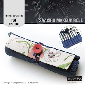May include: A navy blue makeup roll with a floral patterned flap, secured by a red button and braided cord. The roll is designed to hold makeup brushes. The text "SAACIBO MAKEUP ROLL" is visible.