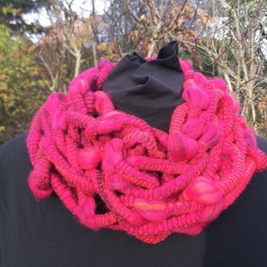 Könnte beinhalten: Handgestrickter Loop-Schal in leuchtendem Pink. Das Design mit grobem Strick weist dickes, strukturiertes Garn mit einer Mischung aus Rosa- und Orangetönen auf. Der Schal ist um ein schwarzes Kleidungsstück gewickelt und zeigt seine kräftige Farbe und einzigartige Textur.