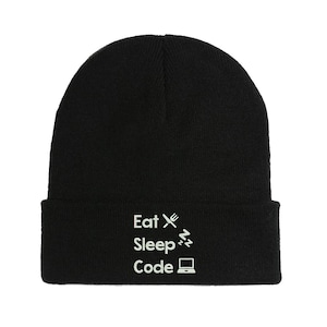 Op de afbeelding: Zwarte gebreide beanie met witte geborduurde tekst die "Eat Sleep Code" zegt met een vork, slapende Z's en een laptop-icoon.