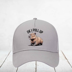 May include: A light gray baseball cap featuring a capybara illustration. The capybara is brown and tan, with the text "OK I PULL UP" above it in black. The cap has a curved brim and a rounded crown.