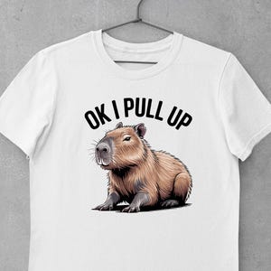 May include: White t-shirt featuring a capybara illustration. The capybara is depicted in shades of brown and gray, with the text "OK I PULL UP" above it in black. The shirt is hanging on a metal hanger.