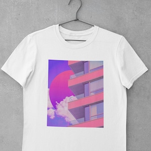 May include: White t-shirt with a graphic print of a pink building with a large pink circle in the sky. The building has multiple windows and balconies.