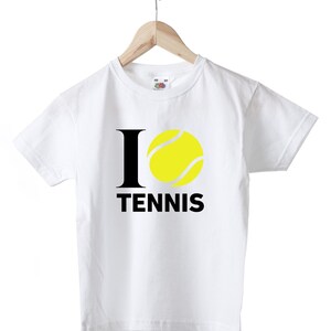 Può includere: T-shirt bianca con la scritta "I ❤️ TENNIS" in nero e una grafica di pallina da tennis gialla. La maglietta è appesa a una gruccia di legno.