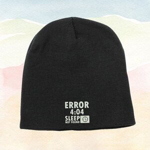 May include: Black beanie with the text "ERROR 4:04 SLEEP NOT FOUND" in white. The beanie is a knit cap, perfect for cold weather. The text includes a clock icon.