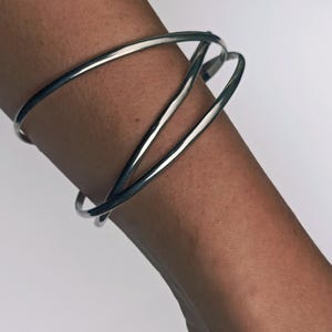 Könnte beinhalten: Ein silberfarbenes Armreif-Armband mit modernem, minimalistischem Design. Das Armband hat drei ineinander verschlungene, gebogene Bänder, die eine offene, geometrische Form bilden. Das polierte Metall reflektiert das Licht und wirkt hell und glänzend. Ca. 6 cm Durchmesser.