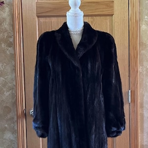 May include: A long black fur coat with a classic collar and a single button closure. The coat is displayed on a white mannequin.