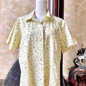 May include: A yellow button-down shirt with a floral pattern of small blue flowers. The short-sleeved shirt is displayed on a mannequin. The shirt has a collar and a single pocket on the chest.