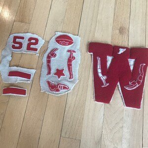 May include: A red felt letter W with white stitching and a red felt football with white stitching. There are also several other red felt patches with white stitching, including a number 52, a star, and two figures.