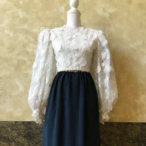 K&ouml;nnte beinhalten: Ein wei&szlig;es Spitzenoberteil mit langen &Auml;rmeln, hohem Kragen und einem marineblauen Rock. Das Oberteil hat einen goldenen G&uuml;rtel um die Taille.