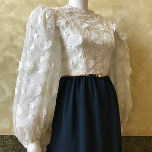 K&ouml;nnte beinhalten: Ein wei&szlig;es Kleid mit einem floralen Spitzenoberteil und langen &Auml;rmeln. Das Kleid hat einen dunkelblauen Rock und einen goldenen G&uuml;rtel.