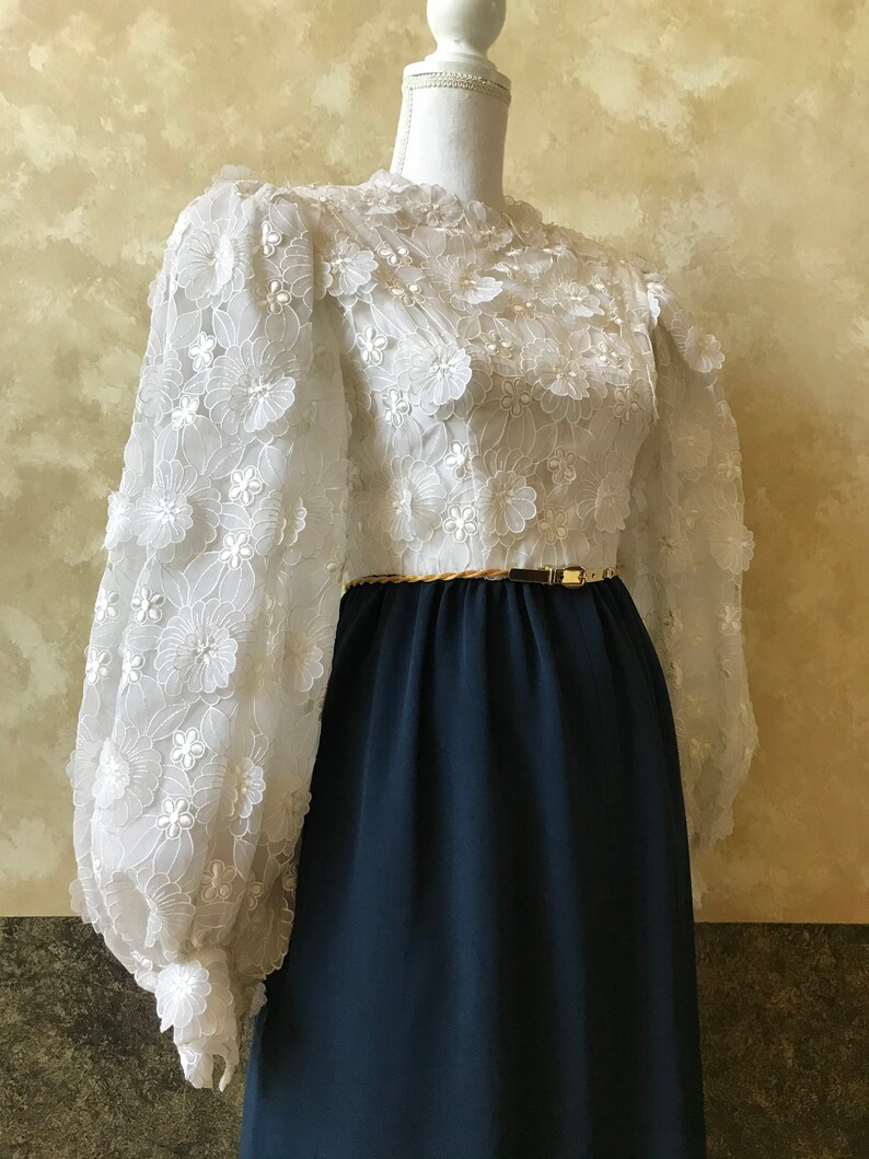 K&ouml;nnte beinhalten: Ein wei&szlig;es Kleid mit einem floralen Spitzenoberteil und langen &Auml;rmeln. Das Kleid hat einen dunkelblauen Rock und einen goldenen G&uuml;rtel.