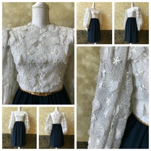 K&ouml;nnte beinhalten: Ein wei&szlig;es Kleid mit einem floralen Spitzenoberteil und einem marineblauen Rock. Das Kleid hat lange &Auml;rmel mit Puff&auml;rmeln und einen goldenen G&uuml;rtel.