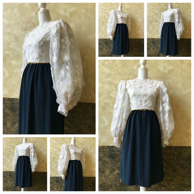 K&ouml;nnte beinhalten: Ein wei&szlig;es Spitzenkleid mit langen, bauschigen &Auml;rmeln und einem dunkelblauen Rock. Das Kleid hat einen goldenen G&uuml;rtel um die Taille.