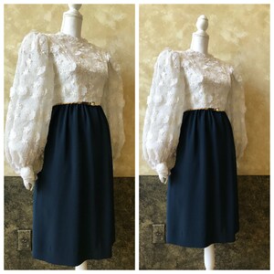 K&ouml;nnte beinhalten: Ein Vintage-Kleid mit einem wei&szlig;en, blumigen Oberteil mit Puff&auml;rmeln und einem dunkelblauen Rock. Das Kleid hat einen goldenen G&uuml;rtel.