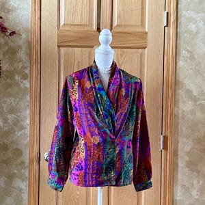 May include: A colorful, long-sleeved blouse with a draped neckline. The blouse features a vibrant pattern of purple, green, orange, and brown. The garment is displayed on a white mannequin against a wooden door background.