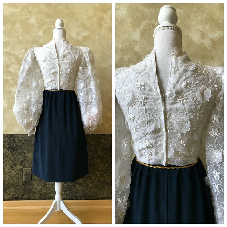 K&ouml;nnte beinhalten: Ein wei&szlig;es Kleid mit einem dunkelblauen Rock und einem goldenen G&uuml;rtel. Das Kleid hat ein Spitzenoberteil mit einem Blumenmuster und langen &Auml;rmeln. Der Rock ist knielang und hat ein schlichtes Design.