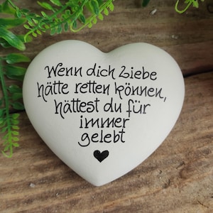 Könnte beinhalten: Weißer herzförmiger Stein mit dem Text "Wenn dich Liebe hätte retten können, hättest du für immer gelebt" in schwarzer Schrift. Ein kleines schwarzes Herz befindet sich am unteren Rand des Steins.