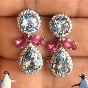 Puede incluir: Un par de pendientes colgantes con una piedra preciosa de topacio azul en forma de lágrima y acentos de topacio rosa. Los pendientes están engastados en plata y tienen un halo de diamantes alrededor del topacio azul.
