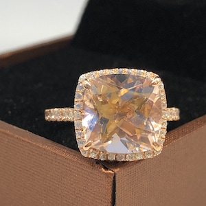 Puede incluir: Un anillo de compromiso de oro rosa con una gran piedra preciosa cuadrada de color rosa claro rodeada por un halo de diamantes más pequeños. El anillo está en una caja de anillo de terciopelo marrón.