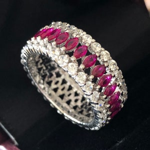 Puede incluir: Un anillo de banda de plata con una fila de rubíes de forma marquesa engastados en pavé. El anillo está rodeado por una fila de diamantes redondos más pequeños.