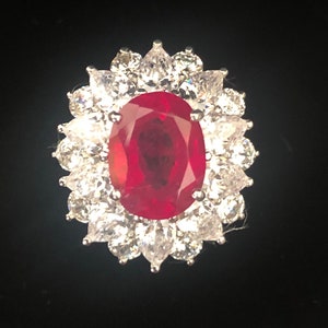 Puede incluir: Un anillo de piedra roja con una gran piedra central de forma ovalada rodeada de piedras blancas más pequeñas.