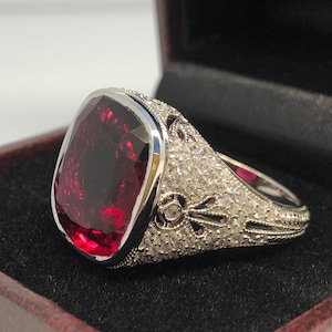 Pode incluir: Um anel de prata com uma grande gema vermelha escura engastada em uma forma quadrada. O anel é decorado com pequenas gemas brancas ao redor da banda.