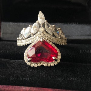 May include: A silver ring with a large heart-shaped ruby surrounded by smaller diamonds. The ring has a crown-like design with marquise-shaped diamonds on the top.