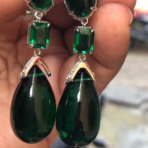 May include: A pair of dangle earrings with green gemstone drops and emerald green accents. The earrings are silver toned and feature a teardrop shaped stone.