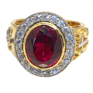 Könnte beinhalten: Ein goldener Ring mit einem großen roten ovalen Edelstein, der von einem Heiligenschein aus kleineren weißen Edelsteinen umgeben ist. Der Ring hat ein detailliertes Design mit zwei Kreuzen an den Seiten.