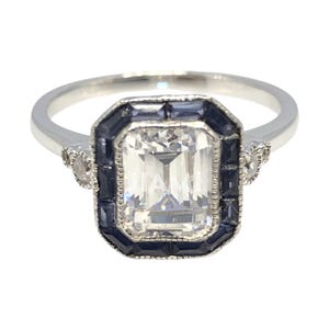 Puede incluir: Un anillo de plata con una gran piedra preciosa transparente de talla esmeralda rodeada por un halo de zafiros azules oscuros más pequeños. El anillo tiene dos piedras preciosas transparentes más pequeñas a cada lado de la piedra principal.