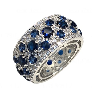 Könnte beinhalten: Ein breiter, silberfarbener Ring mit dunkelblauen Edelsteinen und kleineren klaren Steinen. Das Design des Rings zeigt ein Muster aus dunkelblauen Edelsteinen, die in einem gitterartigen Muster angeordnet sind und von kleineren, klaren Steinen umgeben sind. Ein auffälliges Schmuckstück.
