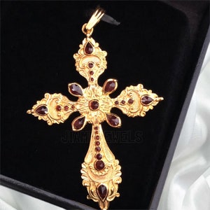 May include: A gold cross pendant with intricate detailing and red gemstones. The cross is set against a black background.