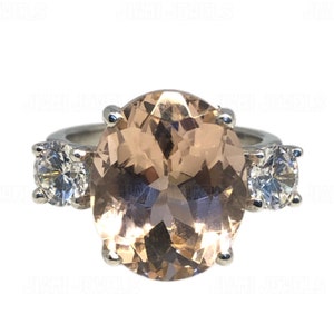 Puede incluir: Un anillo con una piedra preciosa grande, ovalada, de color melocotón rodeada de dos piedras preciosas redondas más pequeñas y transparentes.