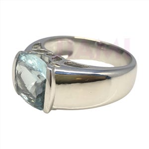 Può includere: Un anello d'argento con una grande pietra preziosa azzurra. La pietra è rettangolare con angoli arrotondati. La fascia dell'anello è spessa e lucida, riflettendo la luce. L'incastonatura è semplice.