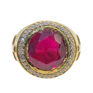 Könnte beinhalten: Ein goldener Ring mit einem großen, ovalen, roten Edelstein, der von einem Heiligenschein aus kleineren, weißen Edelsteinen umgeben ist.