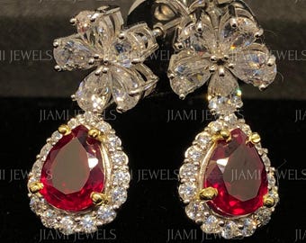 May include: A pair of drop earrings featuring a clear, flower-shaped top set with white stones, connected to a pear-shaped, deep red centre stone surrounded by a halo of small white stones. The metal setting is silver-toned, and the text JIAMI JEWELS is visible.