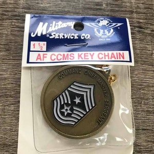 May include: A gold-colored keychain with a round metal charm featuring the text "COMMAND CHIEF MASTER SERGEANT" and a black and white insignia. The keychain is packaged in a clear plastic bag with a blue and white card that reads "Military Service Co. AF CCMS KEY CHAIN 1 1/2"