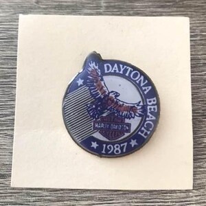 May include: A vintage Harley-Davidson pin featuring a soaring eagle design. The pin is circular with a blue and white background, the words "Daytona Beach" and "1987" are visible. The pin is on a square piece of paper.