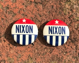 May include: Two red, white, and blue political campaign buttons with a white star and the word "Nixon" in blue letters.