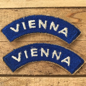 May include: Two blue and white embroidered patches with the word "VIENNA" on them. The patches are curved and have a tan border.