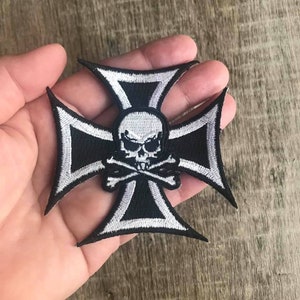 May include: Black and white embroidered iron-on patch featuring a skull and crossbones design in the center of a silver and black iron cross.