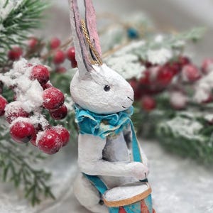 May include: A white paper mache bunny rabbit ornament playing a drum. The bunny is wearing a blue and gold collar and a blue ribbon. The drum is red, blue, and gold.