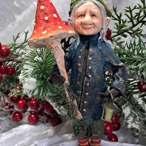 May include: A whimsical figurine of an elf holding a large red mushroom with white spots. The elf wears a blue coat, green hat, and holds a lantern. The background includes red berries and evergreen branches, creating a festive, fairytale theme.
