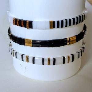 Peut inclure: Trois bracelets superposés composés de perles rectangulaires noires, blanches et dorées métallisées. Le bracelet supérieur présente des segments alternés noirs et blancs avec des accents dorés. Le bracelet central est principalement noir avec des entretoises dorées. Le bracelet inférieur présente des perles blanches avec des accents noirs et dorés.