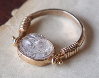 May include: A gold ring featuring a clear, faceted gemstone set in a bezel setting. The ring is crafted from twisted wire and has a simple design.