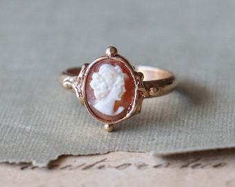 Pode incluir: Um anel de ouro com uma camafeu que apresenta o perfil de uma mulher em branco sobre um fundo marrom.