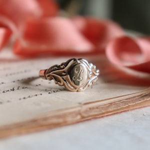 Op de afbeelding: Een goudkleurige ring met een ovale middenstuk met de letter "A" in een scriptlettertype. De ring heeft een sierlijk, filigraan ontwerp rond de ovaal en langs de band. De ring ligt op vintage papier met vervaagde tekst.
