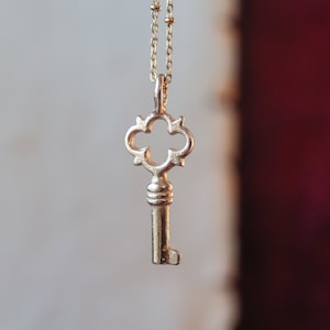 Peut inclure: Un collier avec un pendentif en forme de clé en or et une chaîne délicate. La clé a un design décoratif et orné avec une tête en forme de trèfle.